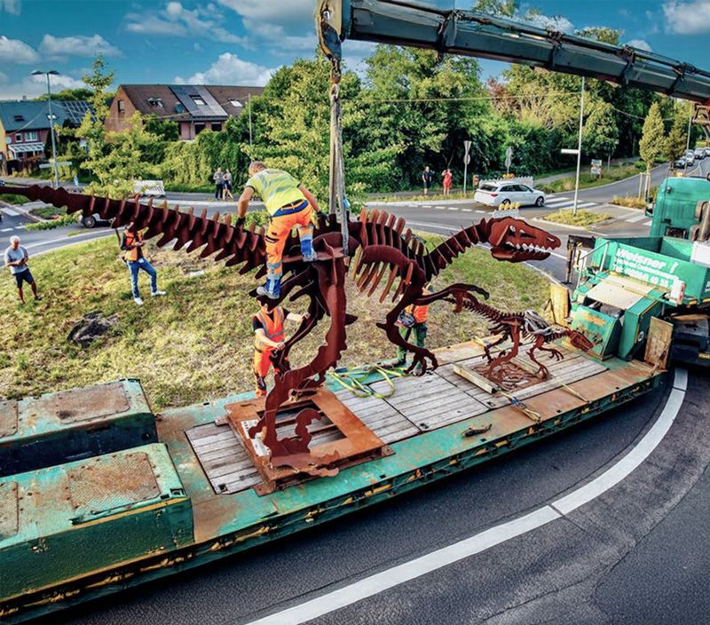 Ein Lastwagen mit Kran hebt das massive Metallskelett eines Dinosauriers auf einen Anhänger, um es an einen neuen Standort zu transportieren.